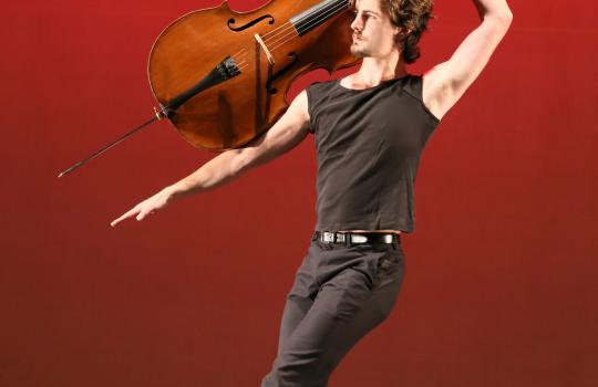 Joseph Teeter on a red stage holding a cello in a dance pose. 