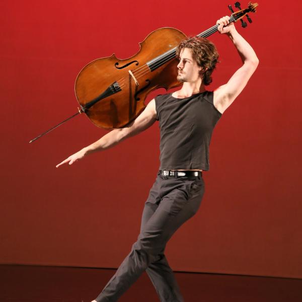 Joseph Teeter on a red stage holding a cello in a dance pose. 